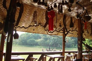 A wooden structure with a thatched roof, outdoor seating, and a view of a lake. - Baan Suan Jantra Home Stay (Chiang Rai)