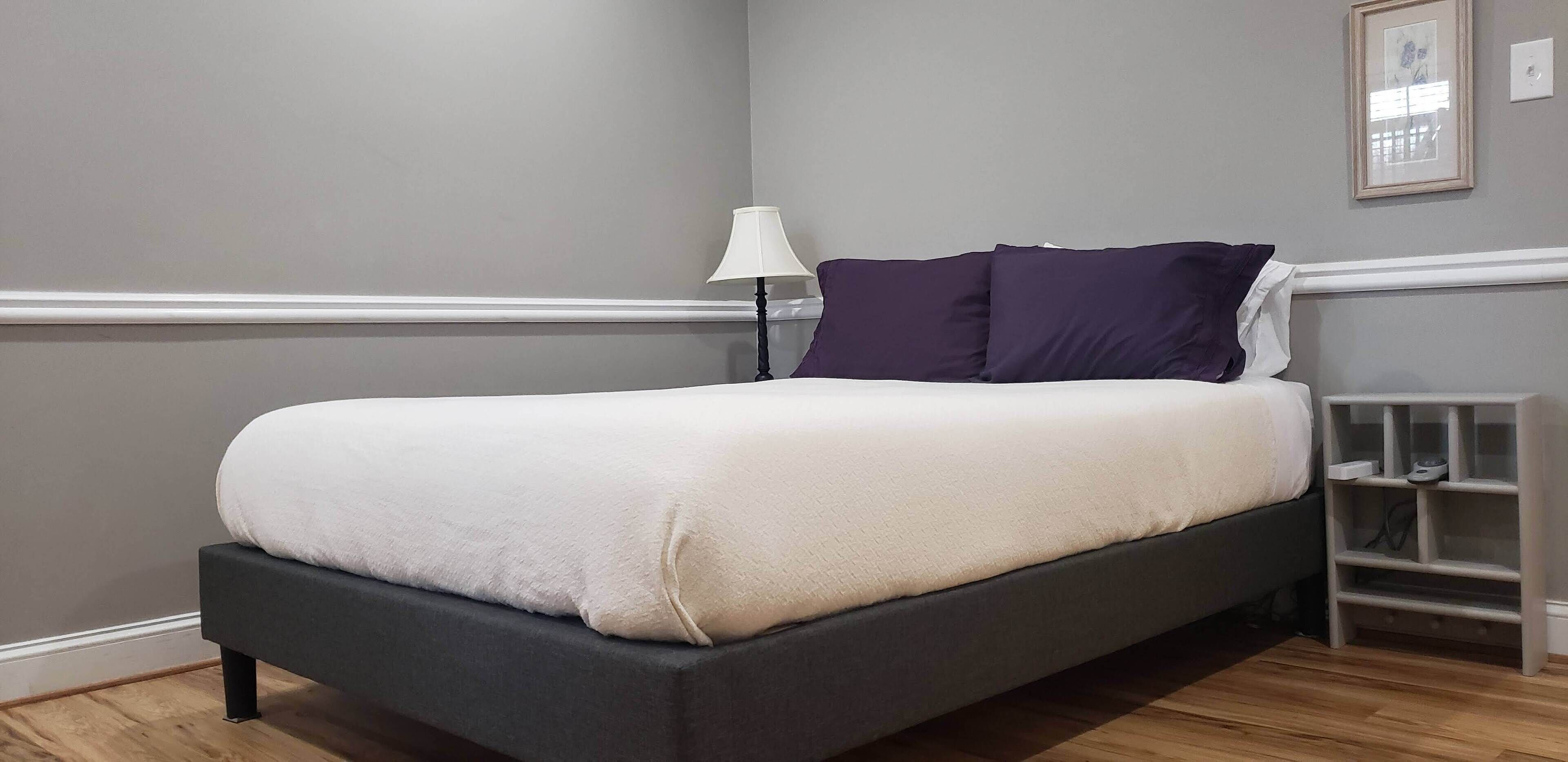 Una cama bien hecha con edredón blanco, cabecero oscuro y mesita de noche con lámpara.