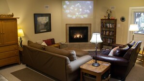 A living room with a fireplace, sofas, a coffee table, a bookshelf, and a television screen displaying a projection.