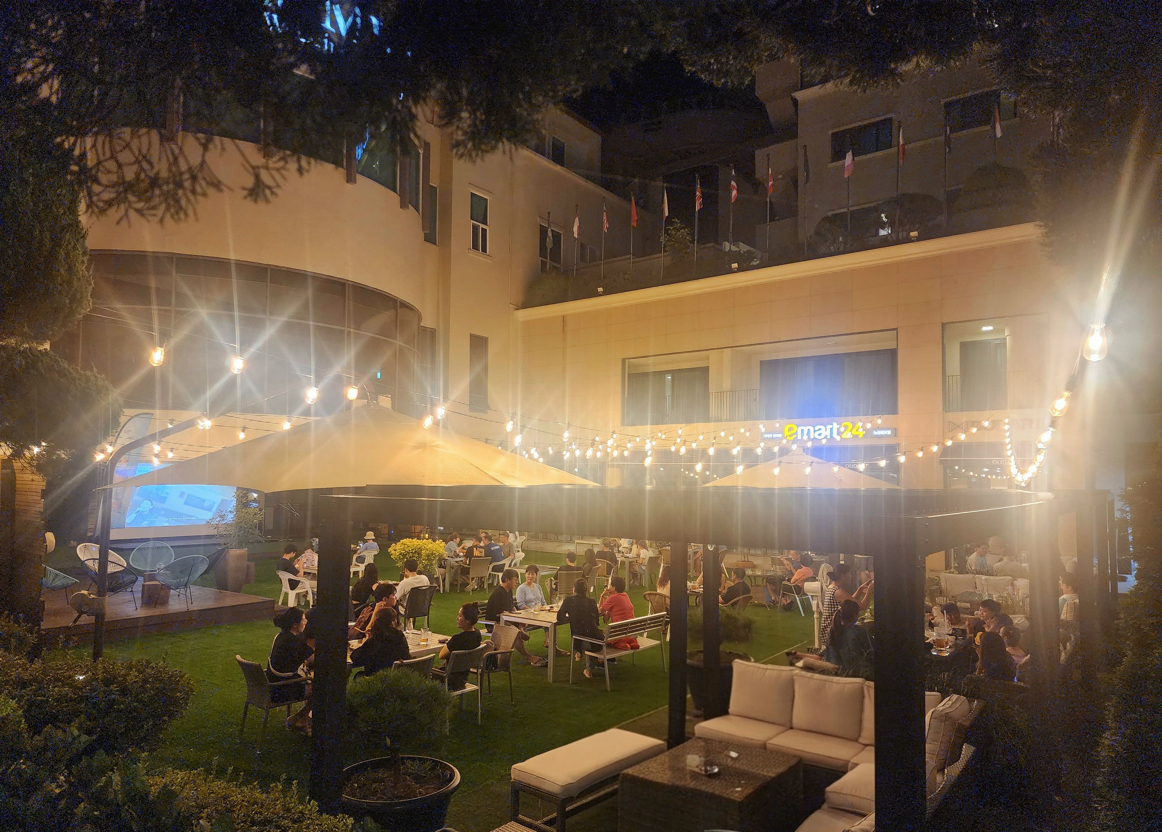 An outdoor dining area with tables and chairs, surrounded by greenery and a building with a visible sign.