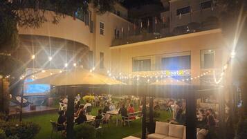 An outdoor dining area with tables and chairs, surrounded by greenery and a building with a visible sign.