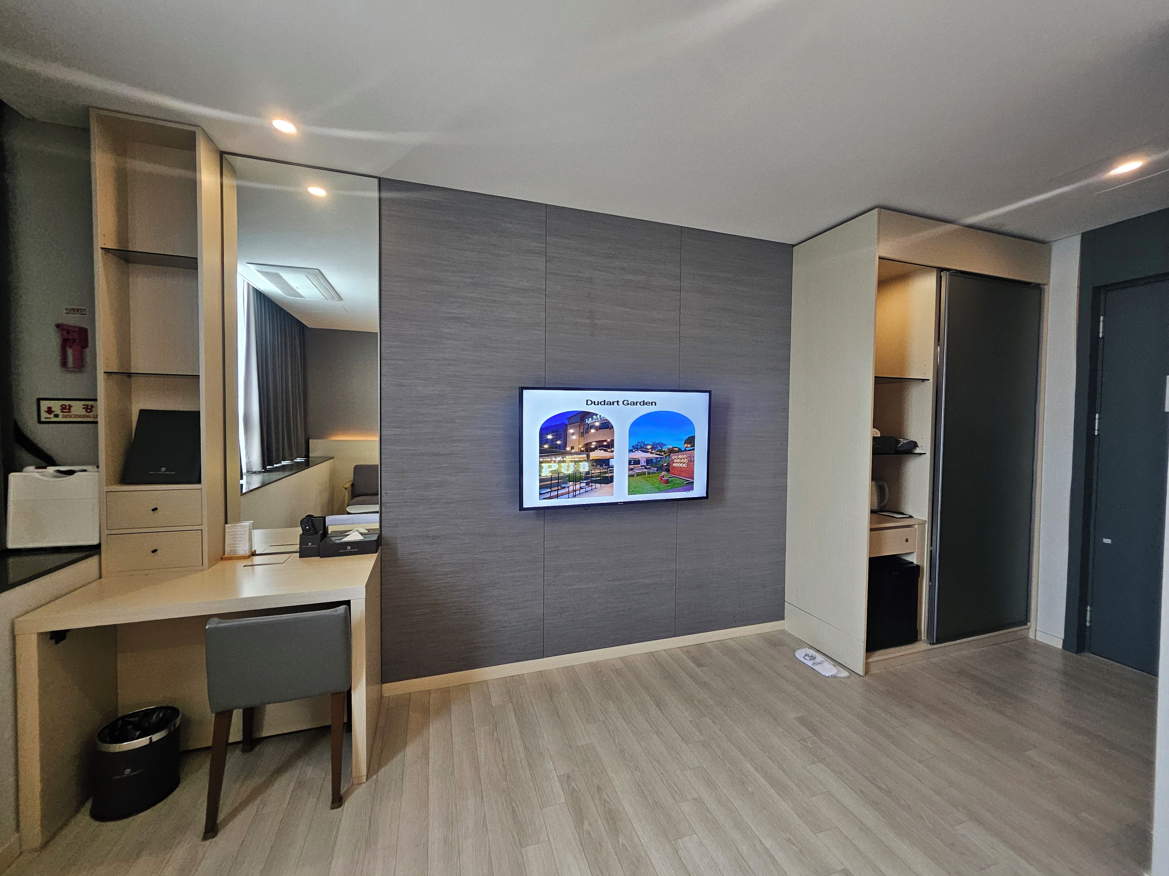 Habitación de hotel moderna con escritorio, silla y televisor montado en la pared mostrando 'Dudart Garden'.