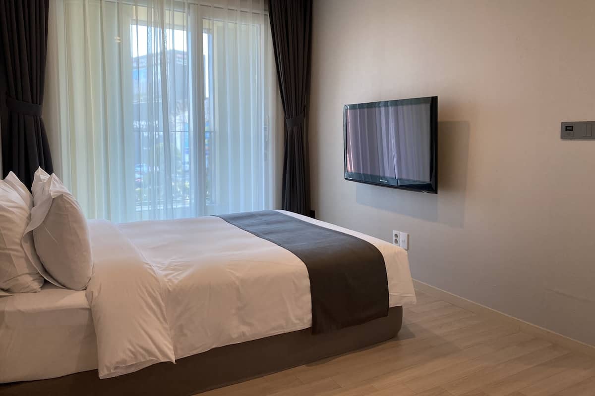 A hotel room with a bed, a television mounted on the wall, and a window with sheer curtains.