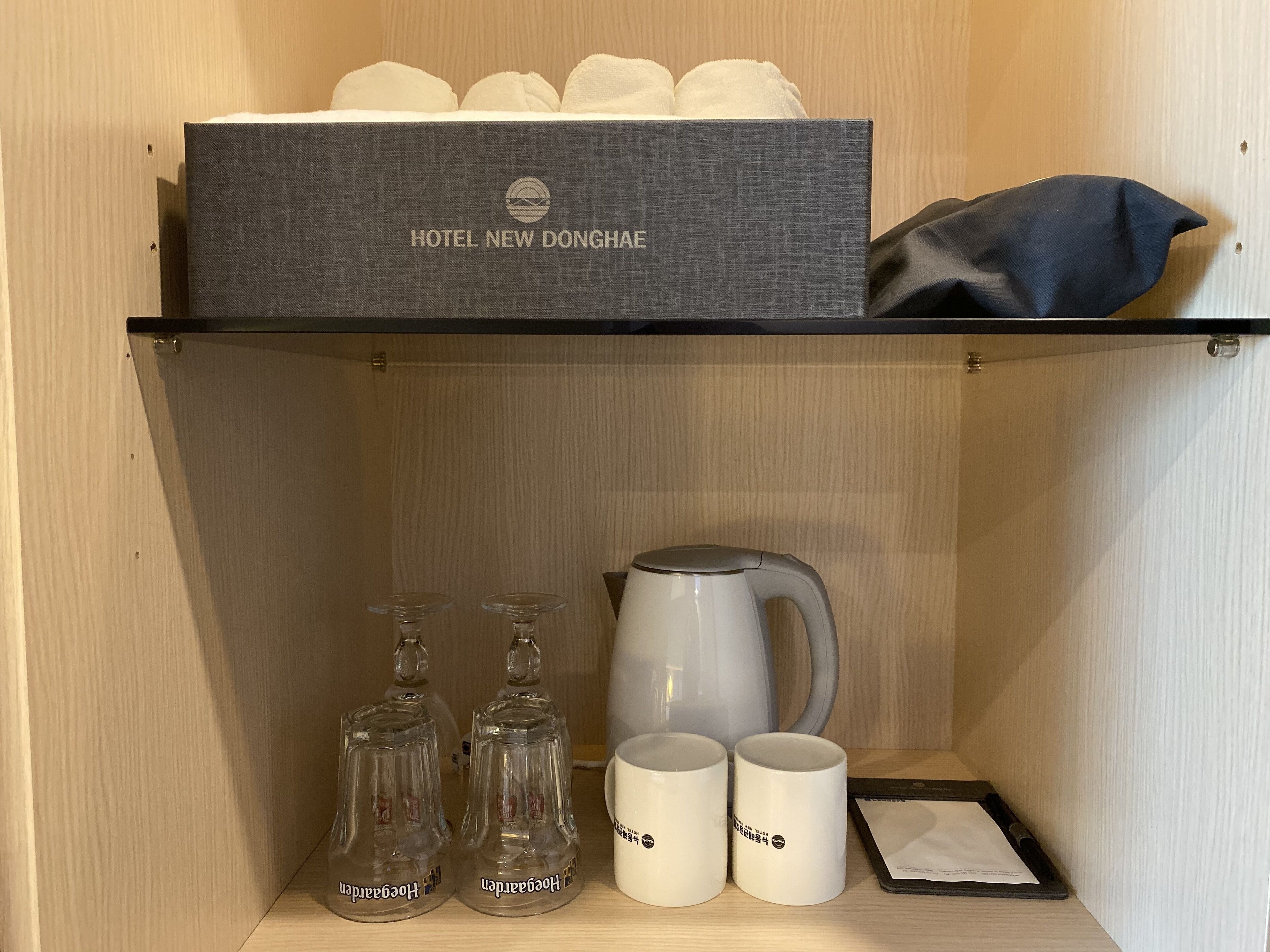 Un minibar de hotel con una caja etiquetada como "HOTEL NEW DONGHAE", una tetera, dos vasos y dos tazas.