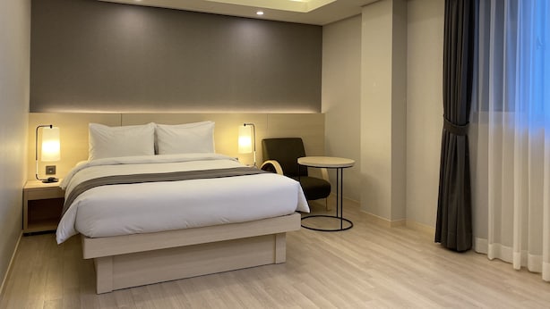 Habitación de hotel moderna con una cama grande, lámparas de noche, una mesita redonda y vistas al exterior a través de cortinas traslúcidas.