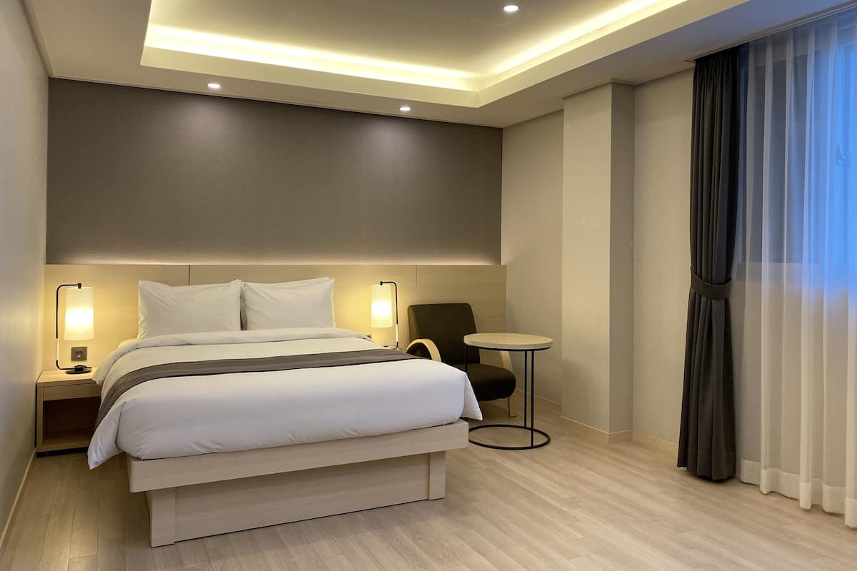 A modern hotel room with a large bed, bedside lamps, a small round table, and a view of the outside through sheer curtains.