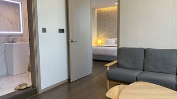 A modern hotel room with a grey sofa, a wooden table, and a bed with a patterned headboard.