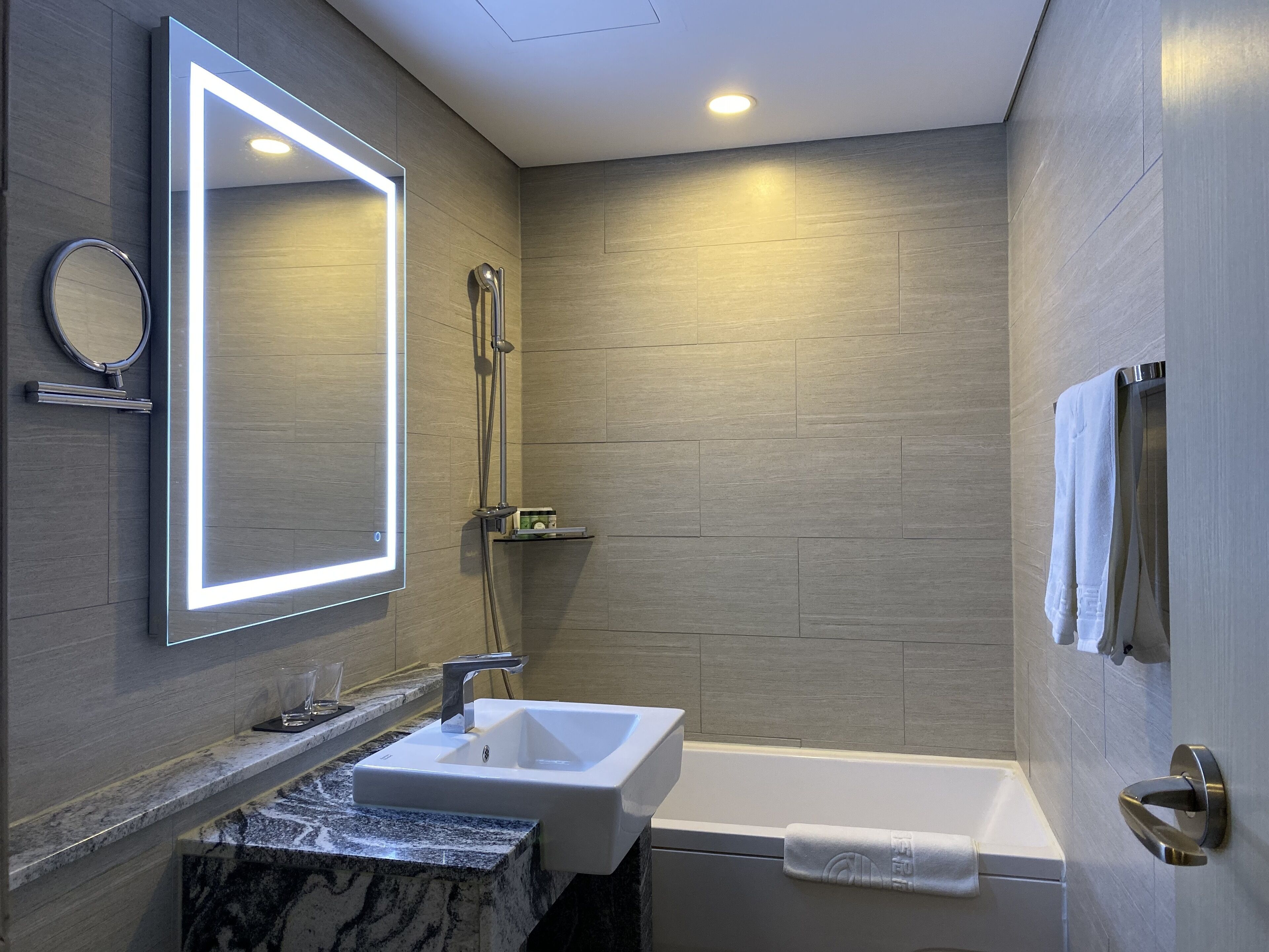 A modern bathroom with a built-in sink, a mirror with illuminated frame, a shower, and a towel rack.