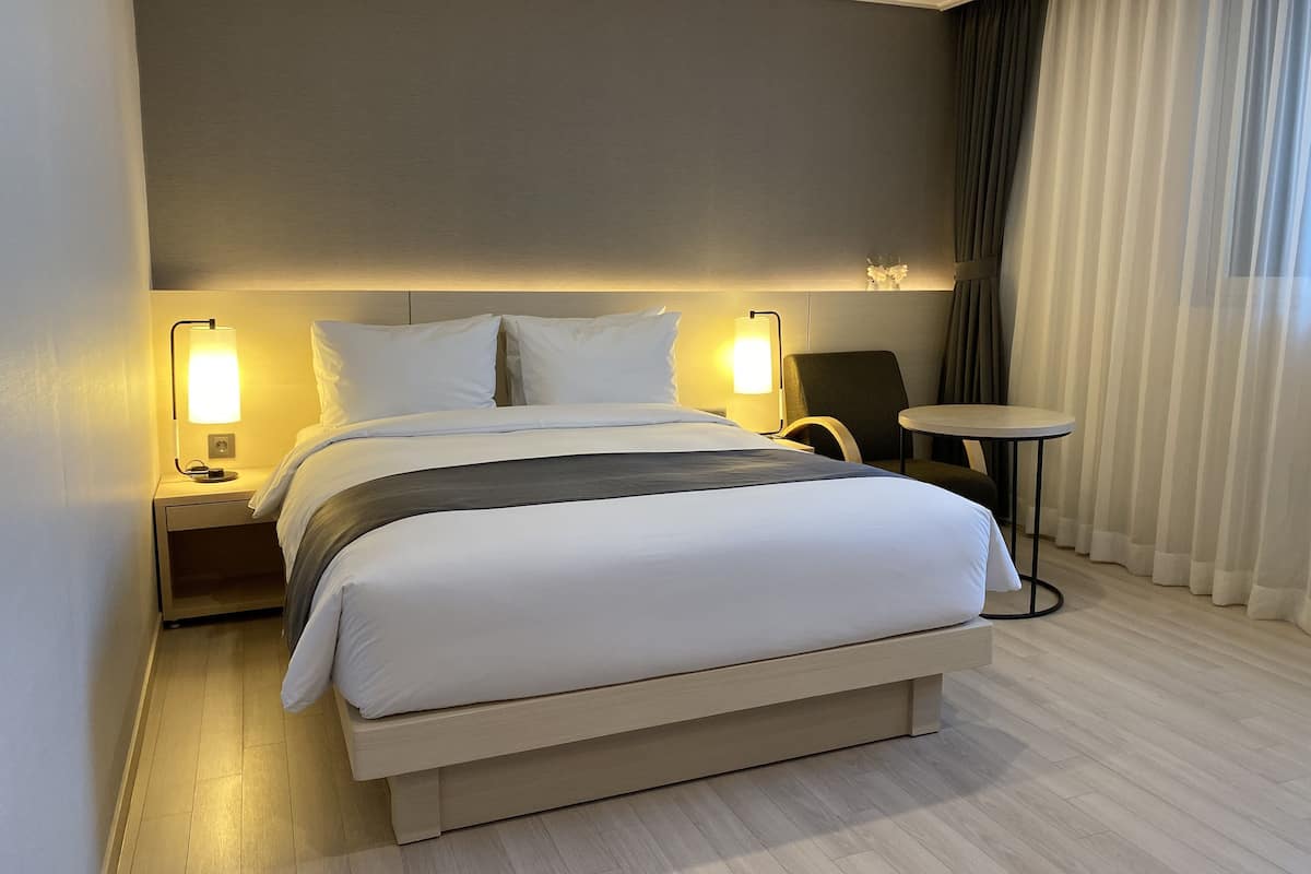 A modern hotel room with a large bed, two bedside lamps, a small round table, and a view of the outside through sheer curtains.