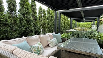 Un área de estar al aire libre con un sofá acolchado y una mesa de vidrio, rodeada de follaje y una estructura cubierta.