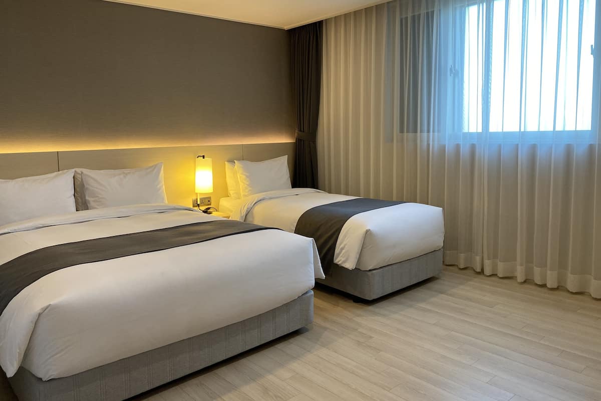A hotel room with two beds, a bedside table with a lamp, a large window with sheer curtains, and a wooden floor.