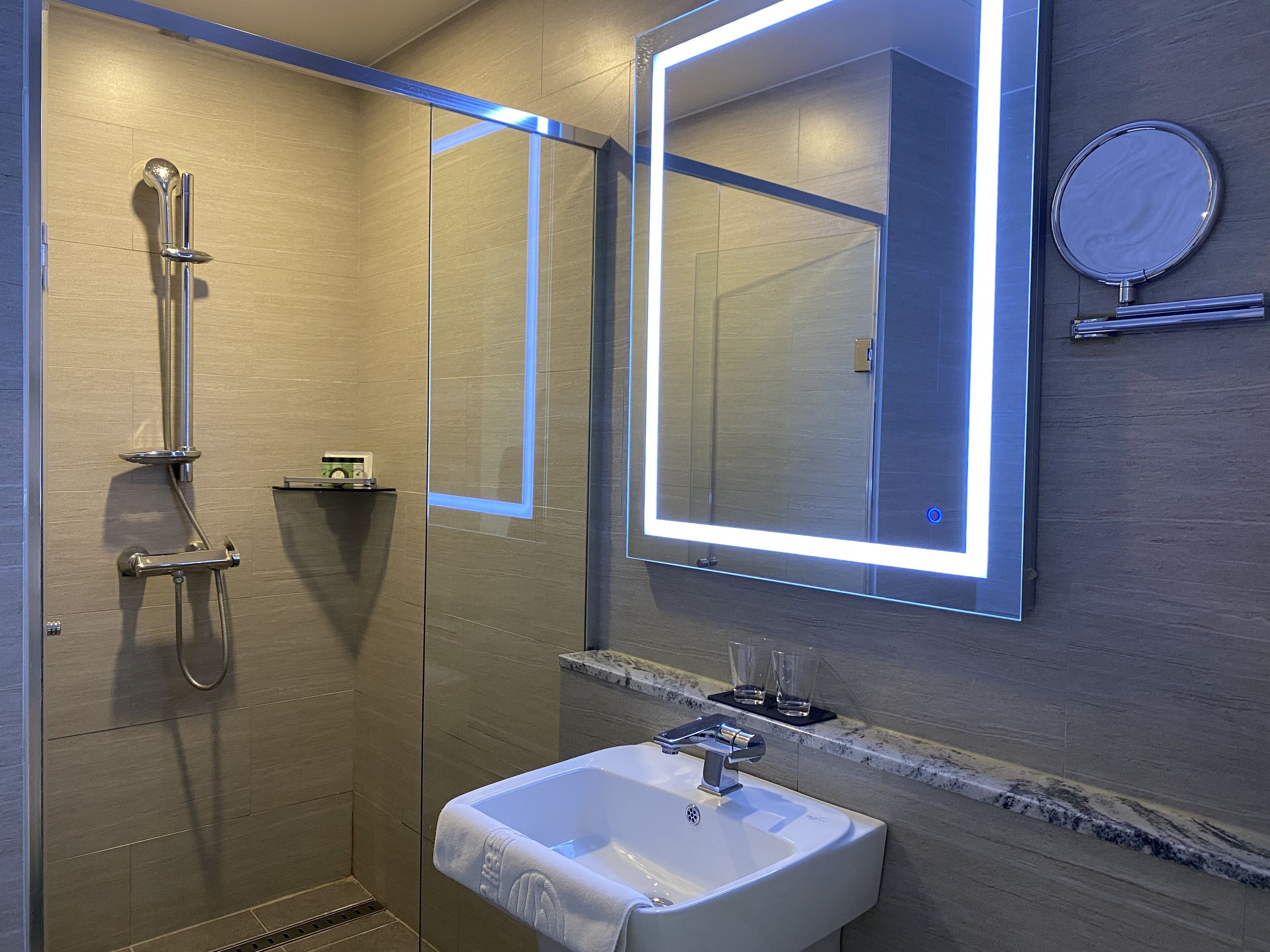 Un baño moderno con ducha acristalada, lavamanos blanco e iluminación integrada en el espejo.