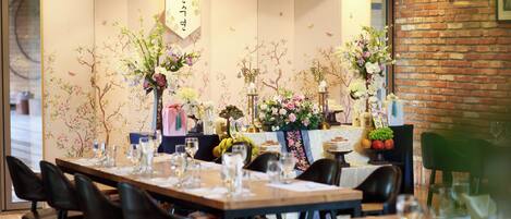 A restaurant interior with a long dining table set for a meal, chairs, and a decorative backdrop with floral arrangements.