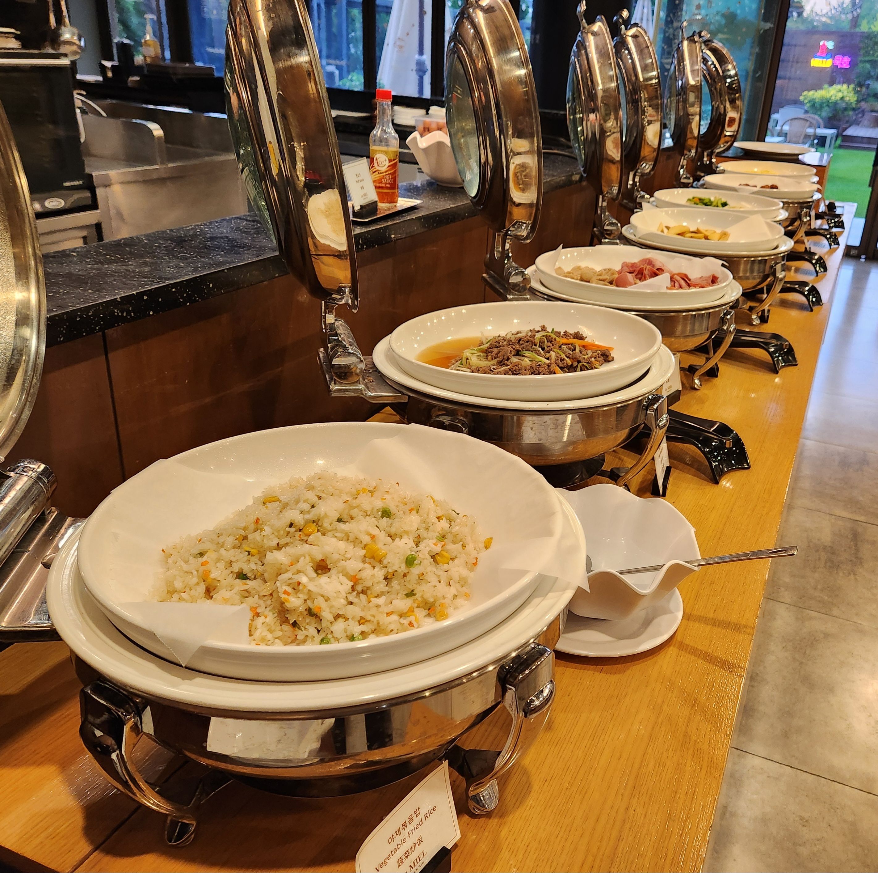 Tarjolla on runsas buffet, jossa on erilaisia ruokia suurissa kulhoissa, mausteita ja kastikkeita erillisessä pisteessä, ja lisäksi avautuu näkymä ulos.