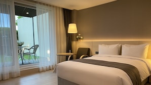 Habitación de hotel con una cama grande, una mesita pequeña, una silla y vistas al exterior a través de cortinas translúcidas.