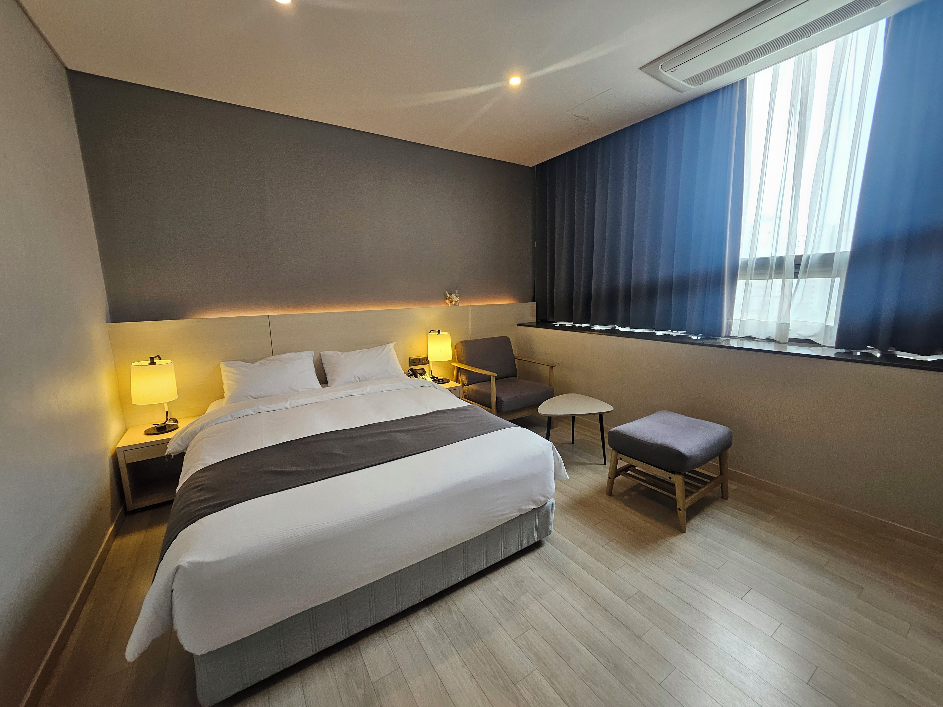 Habitación de hotel con una cama grande, mesitas de noche con lámparas, una pequeña mesa, una silla y una ventana con cortinas.