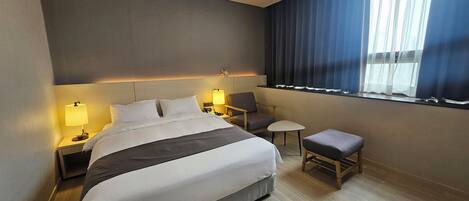 Habitación de hotel con una cama grande, mesitas de noche con lámparas, una pequeña mesa, una silla y una ventana con cortinas.
