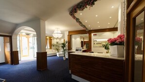 A hotel reception area with a wooden reception desk, a large potted plant, and a chandelier. - Olympic Regina Hotel (Giustino)