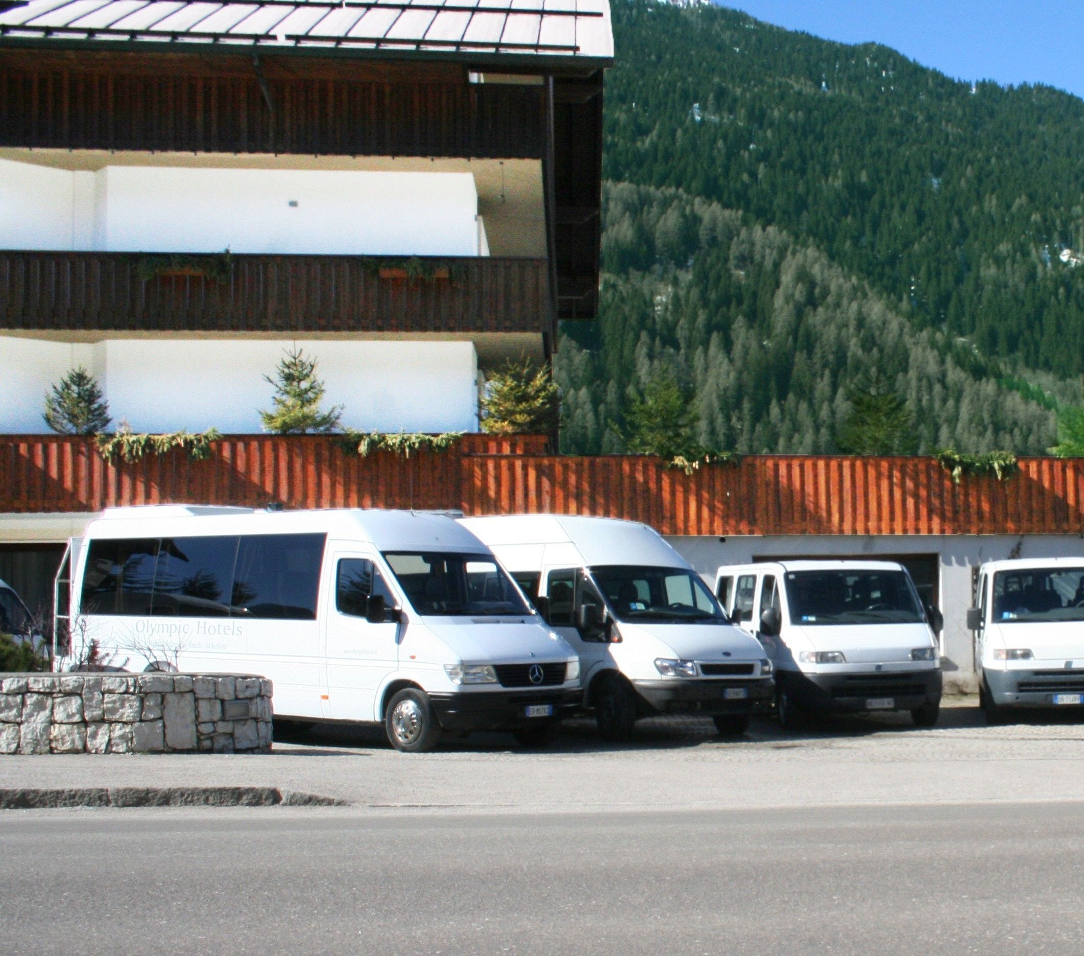 A row of white vans parked in front of a building with a stone wall and a wooden fence.