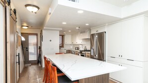 A modern kitchen with a large island, wooden chairs, white cabinets, and stainless steel appliances.