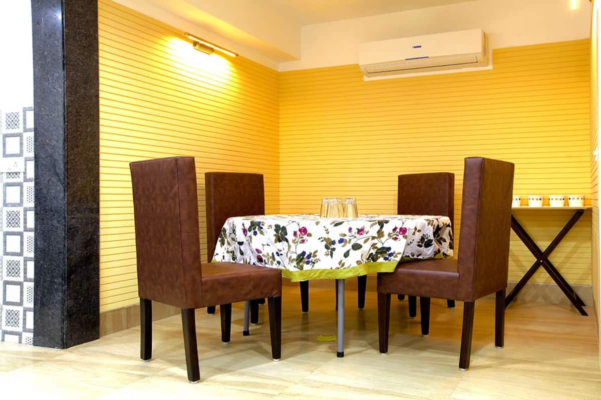 Un comedor con una mesa dispuesta para cuatro personas, rodeada de sillas marrones, frente a una pared amarilla con líneas horizontales.