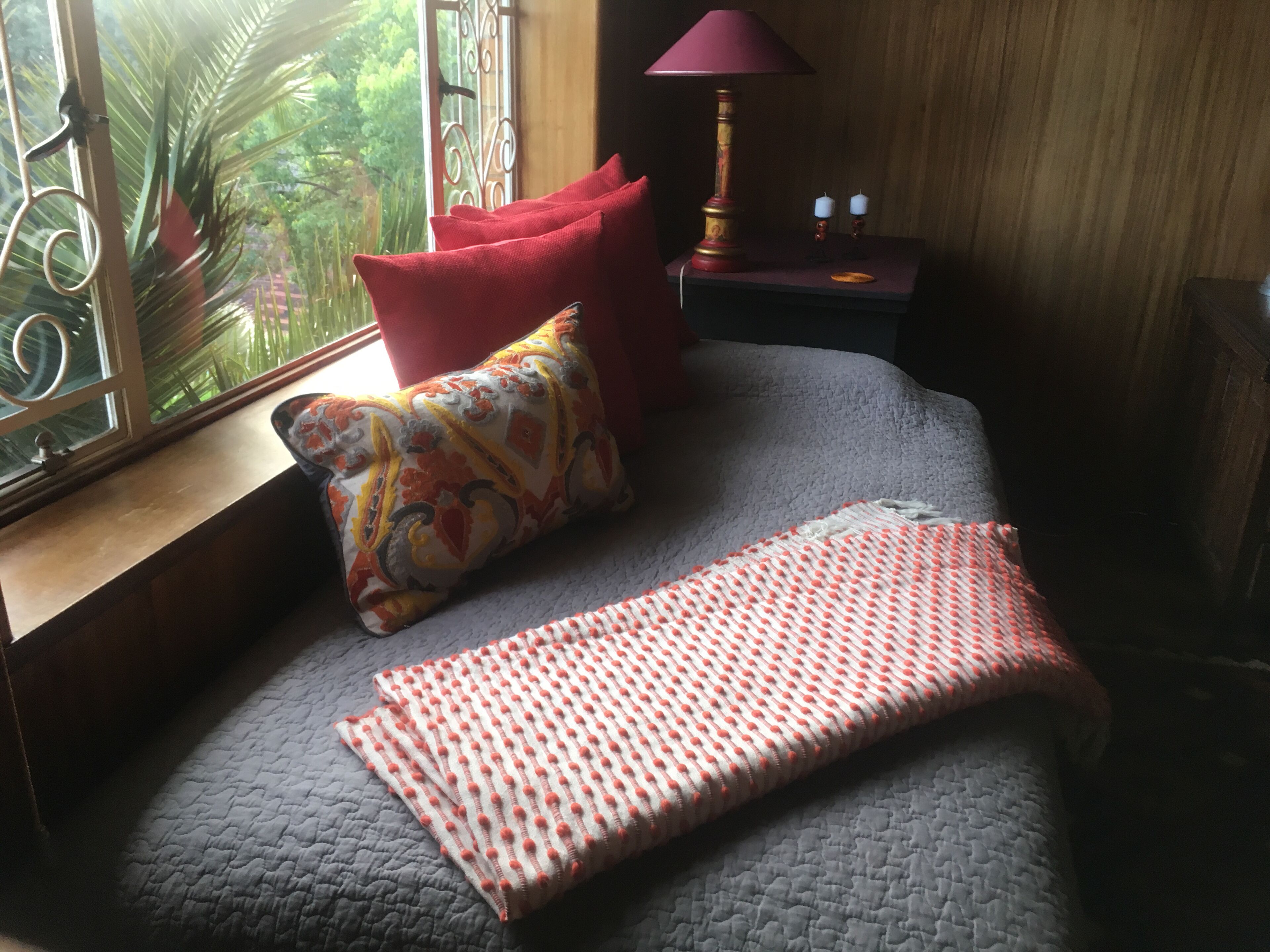 Ein Schlafzimmer mit einem Bett, einem Fenster mit Blick auf Grünflächen, einem Nachttisch mit einer Lampe und einem roten Kissen.