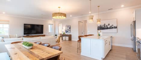 A spacious living room with a wooden dining table, a large flat-screen TV, and a kitchen area with white cabinets and stainless steel appliances.