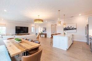 A spacious living room with a wooden dining table, a large flat-screen TV, and a kitchen area with white cabinets and stainless steel appliances.