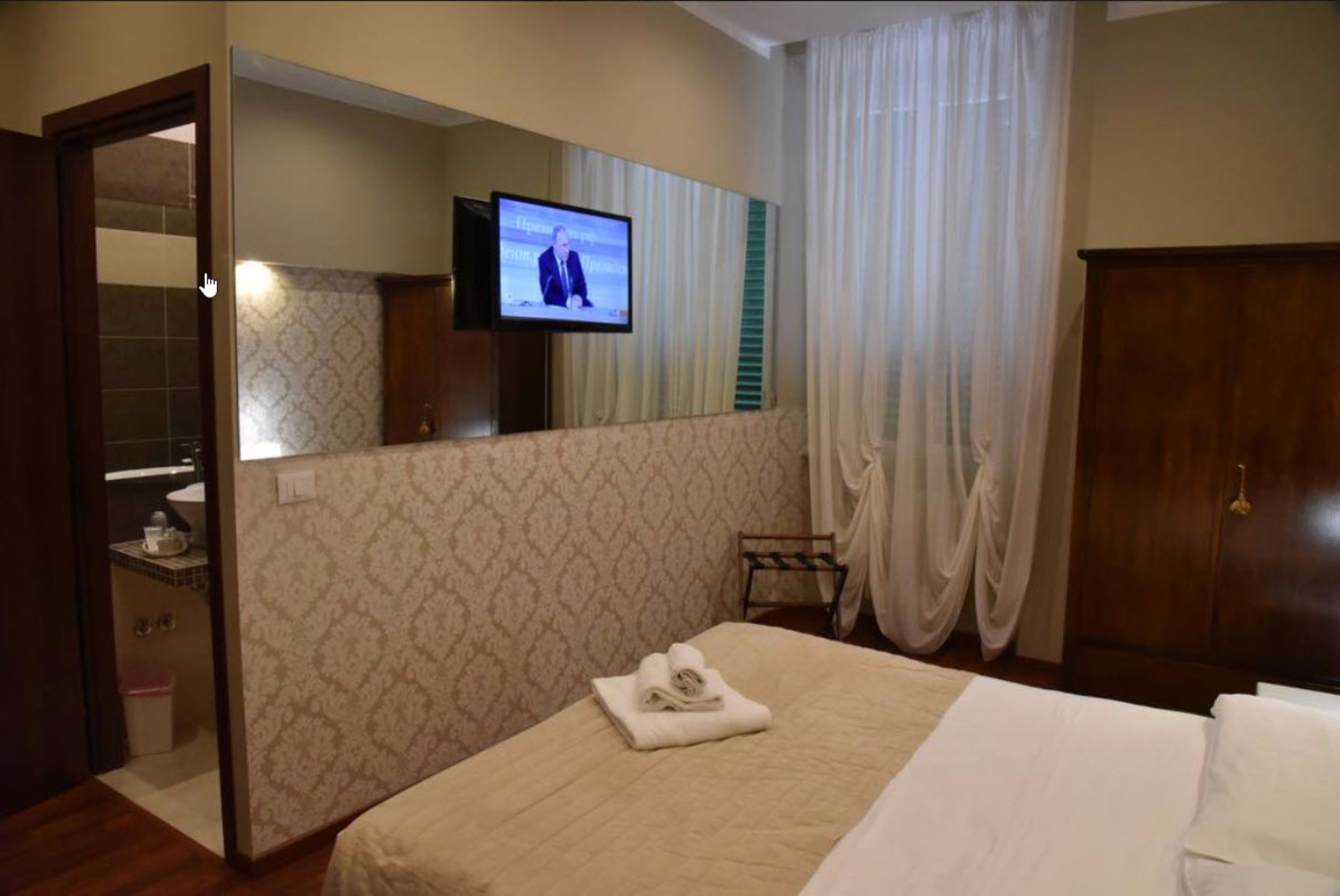 A hotel room with a bed, a television mounted on the wall, a bathroom visible through an open door, and a wooden wardrobe.