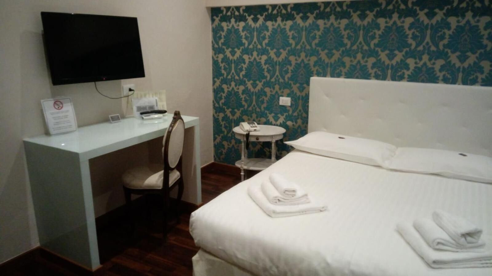A hotel room with a bed, a desk, a chair, a small round table, and a television mounted on the wall.