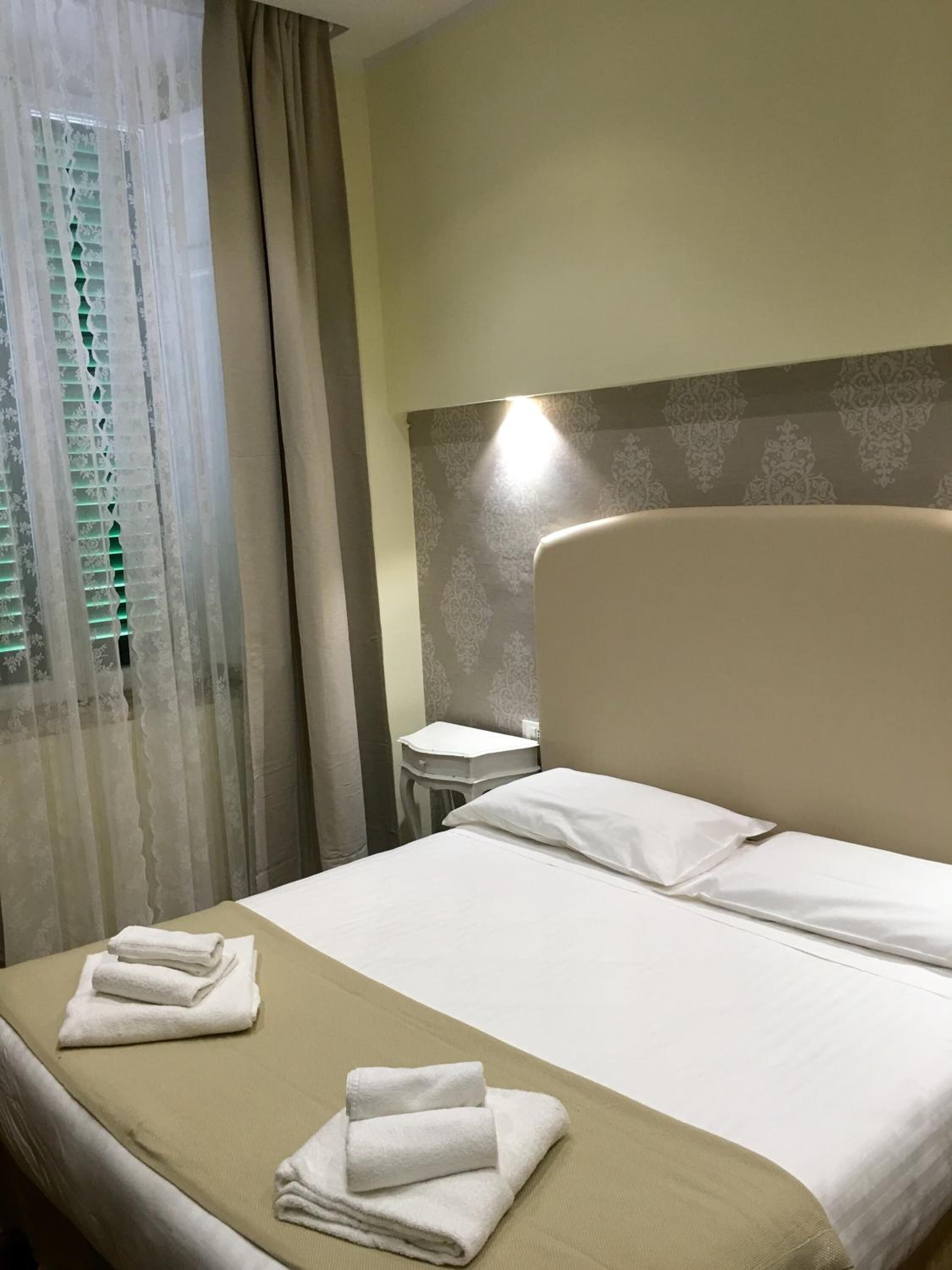 A hotel room with a bed, a nightstand, a window with curtains, and a wall-mounted light.