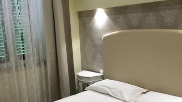 A hotel room with a bed, a nightstand, a window with curtains, and a wall-mounted light.
