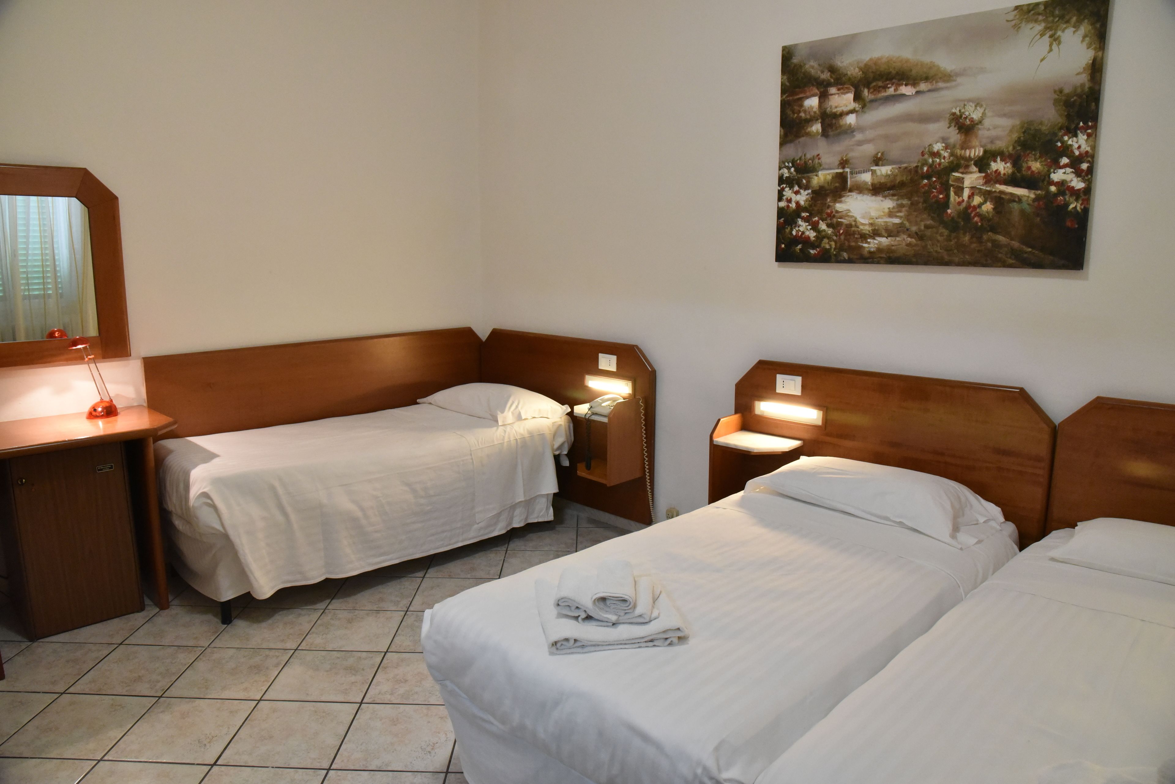 Habitación de hotel con dos camas individuales, cabecero de madera, mesita de noche con lámpara, teléfono y un cuadro en la pared.