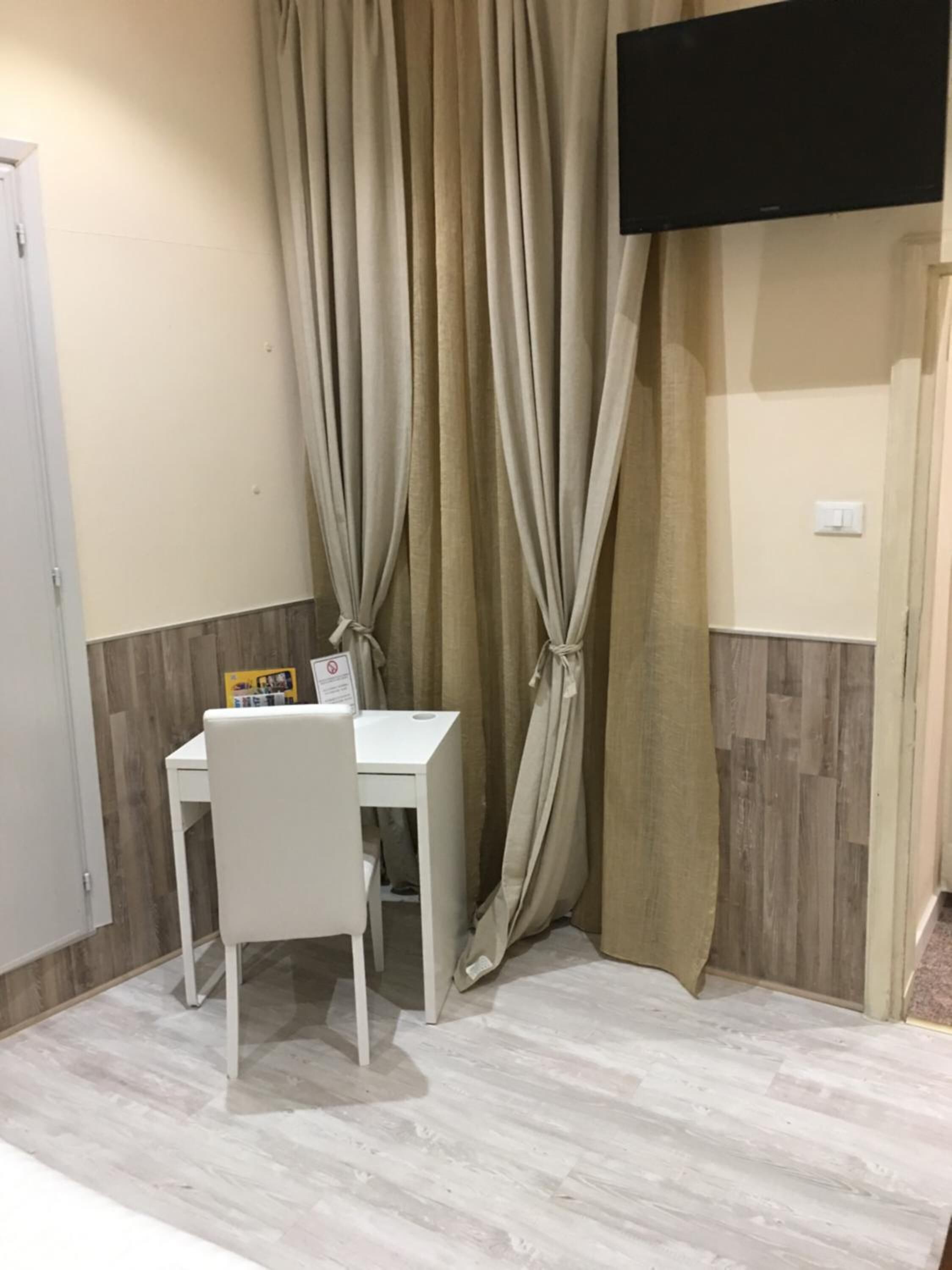 Una stanza piccola con un tavolo e una sedia bianchi, tende color beige, una TV a parete e un pavimento in legno.