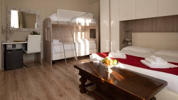 Una habitación de hotel con literas, un escritorio con espejo, un refrigerador pequeño, una mesa de madera con fruta y una colcha roja.