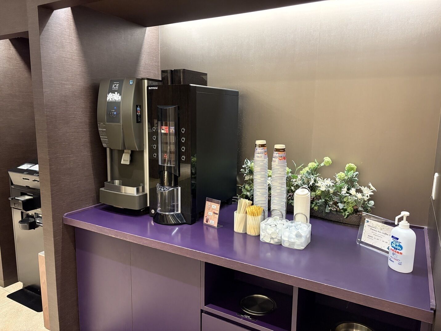 En selvbetjeningskaffestation med kaffemaskine, papkrus og en vanddispenser.