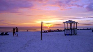 A beach scene with a volleyball net, a gazebo, and people walking.