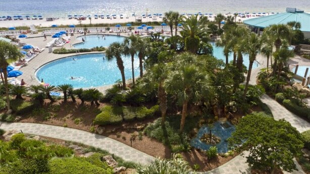 A resort with a large central pool, smaller surrounding pools, palm trees, and a beach area with umbrellas.