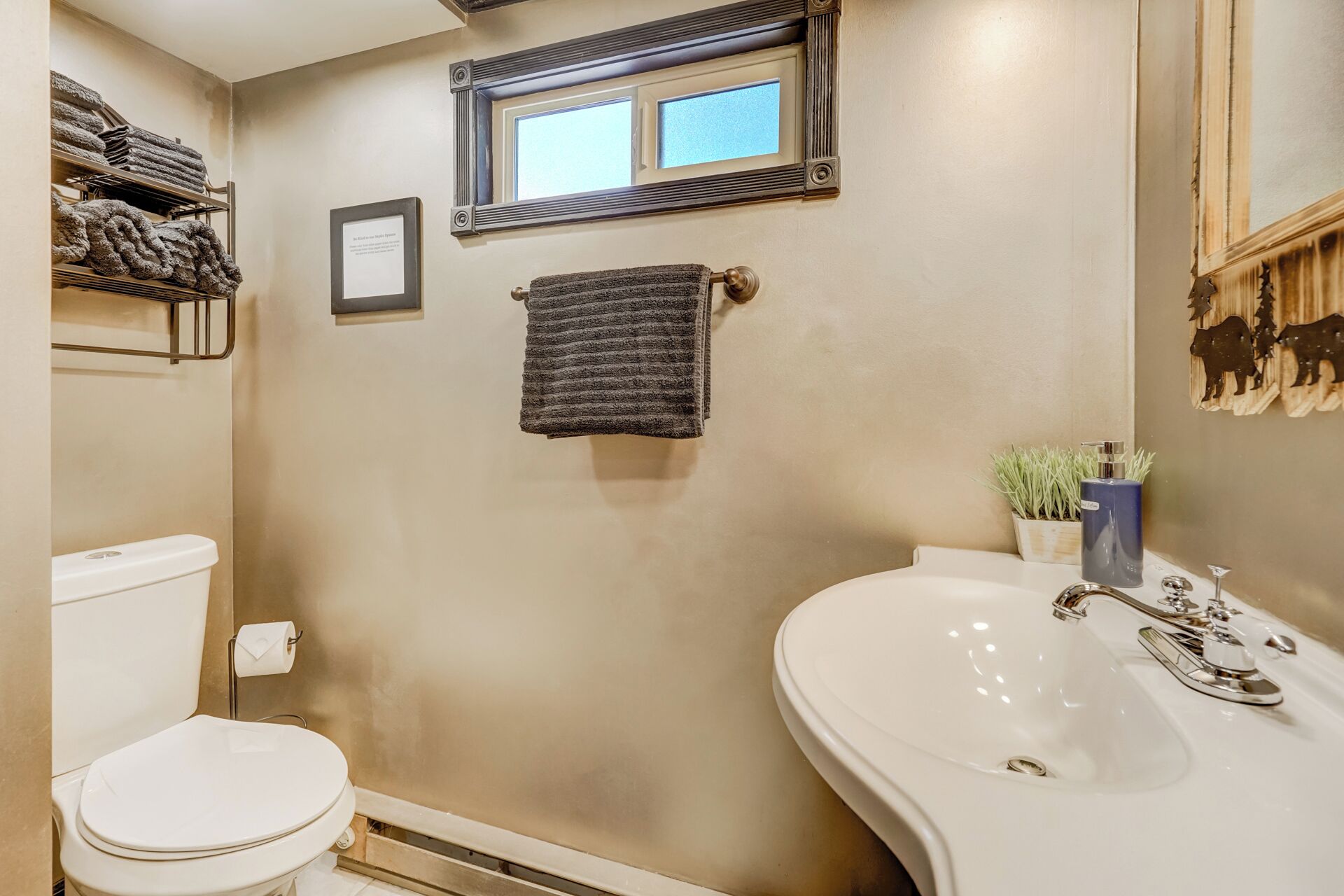 A bathroom with a white toilet, a sink with a mirror, a towel rack with a towel, a framed picture, and a window.