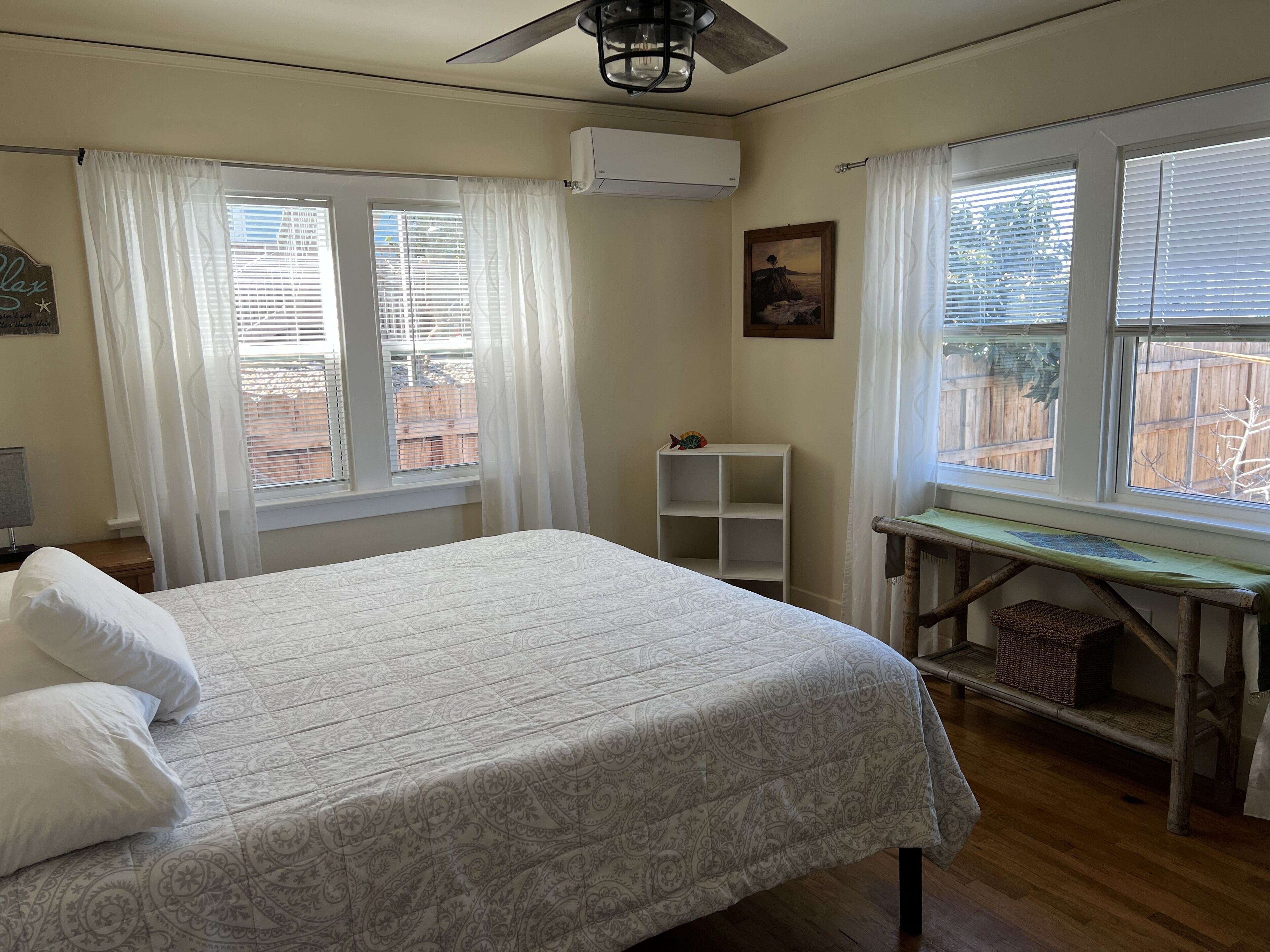 Un dormitorio con una cama, un ventilador de techo y dos ventanas con cortinas blancas.