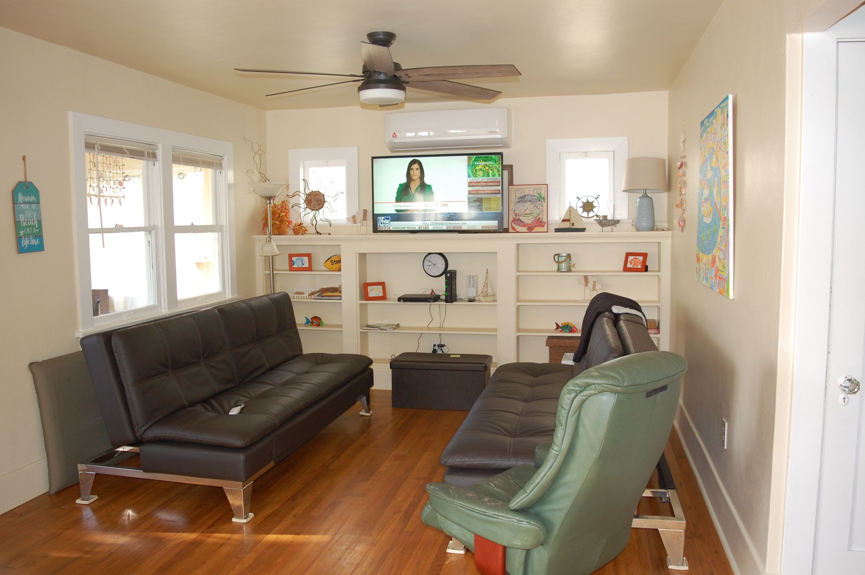 Un salon avec un canapé en cuir marron, un fauteuil vert, un téléviseur fixé au mur et un ventilateur de plafond.