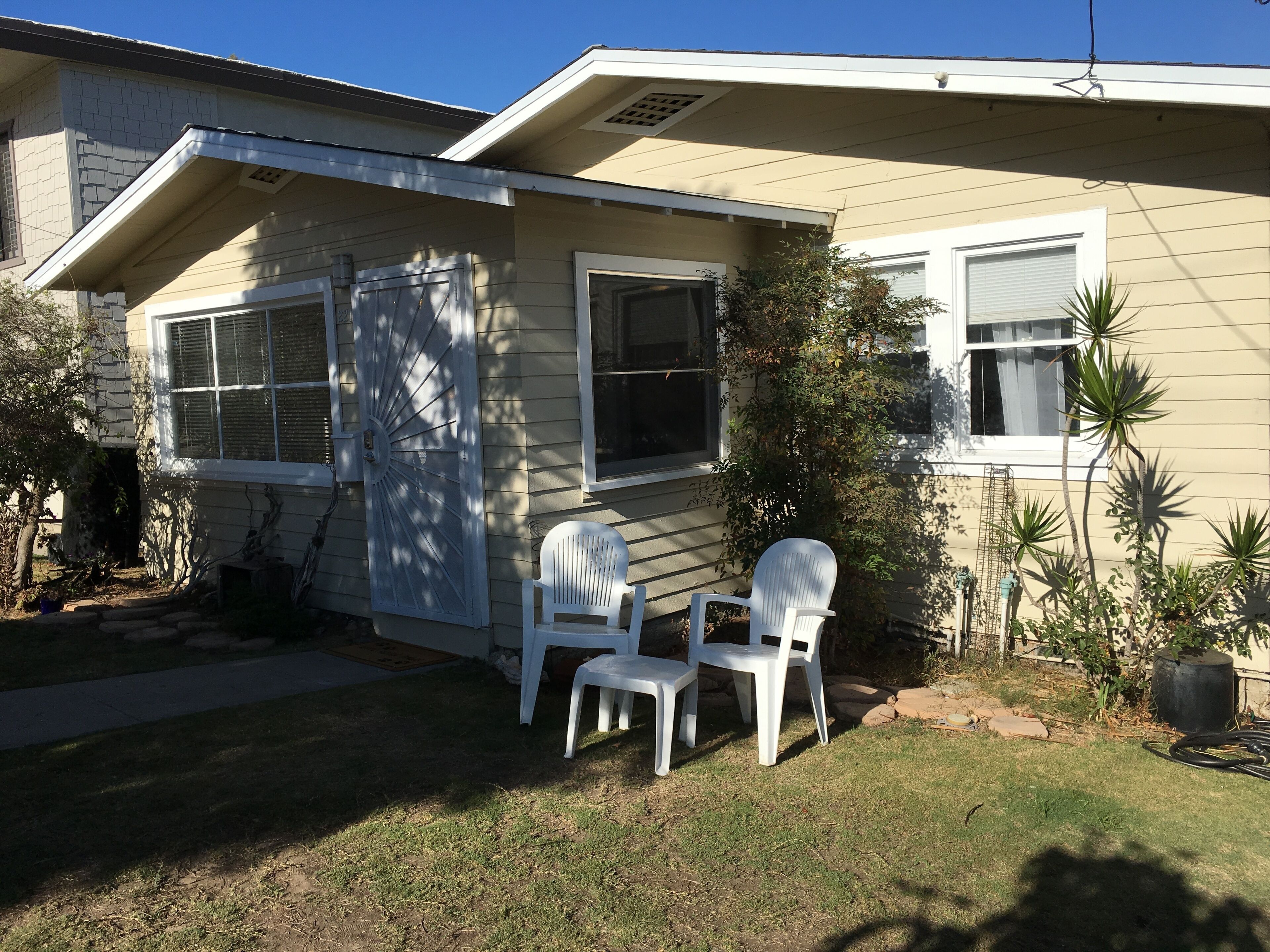 A single-story house with a front porch, two white plastic chairs, and a small garden area with a potted plant.