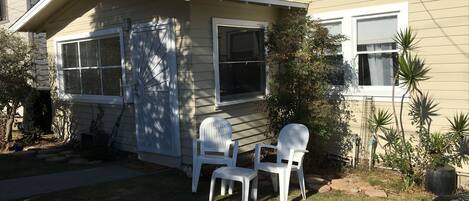 A single-story house with a front porch, two white plastic chairs, and a small garden area with a potted plant.