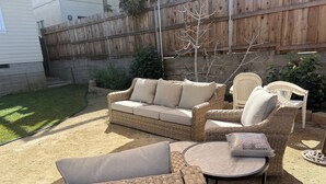 Un patio trasero con mobiliario de mimbre, incluyendo un sofá y sillas, una mesa redonda y un pequeño jardín con cerca de madera y plantas.