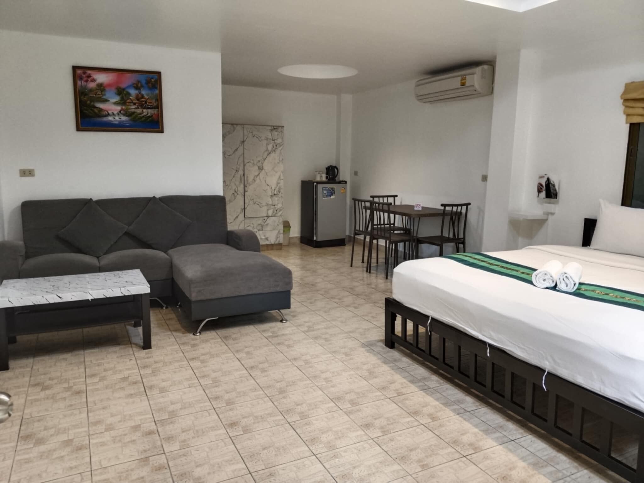 A hotel room with a bed, a sofa, a dining table with chairs, a small refrigerator, and a painting on the wall.