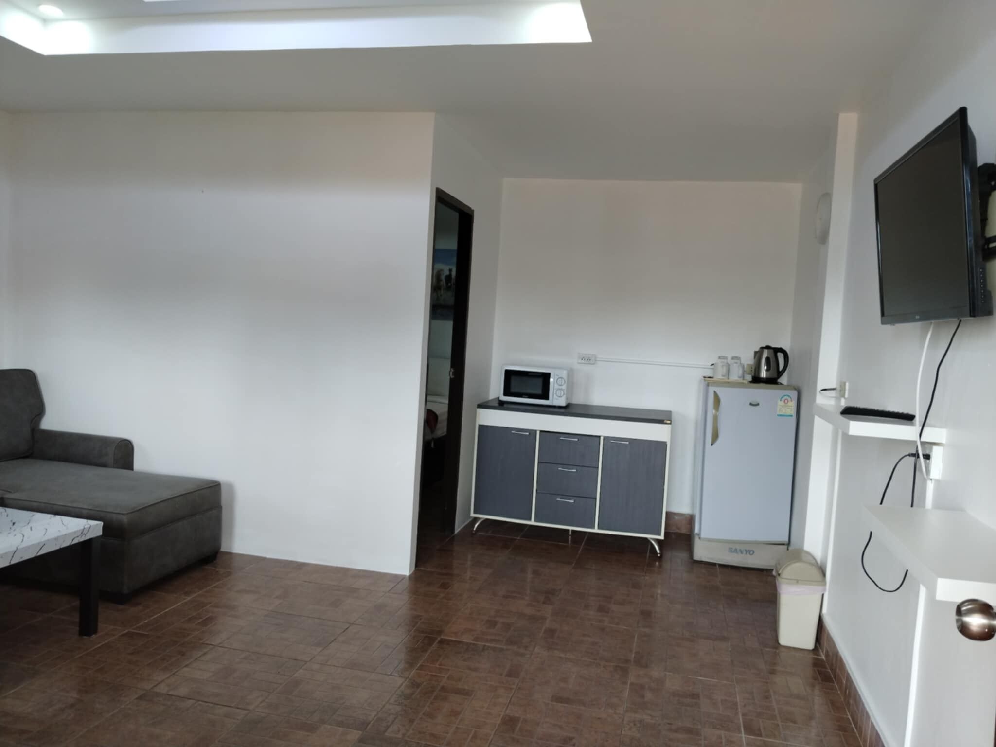 A modern, minimalist room with a grey sofa, a small kitchen area including a microwave and a mini-fridge, and a TV mounted on the wall.