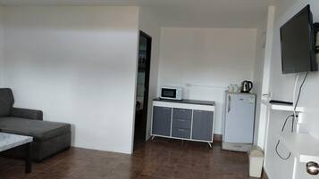 A modern, minimalist room with a grey sofa, a small kitchen area including a microwave and a mini-fridge, and a TV mounted on the wall.