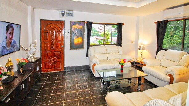 A living room with a sofa, a glass coffee table, and a TV. There is a wooden door and a window with curtains. - Phratamnak Inn (Pattaya)