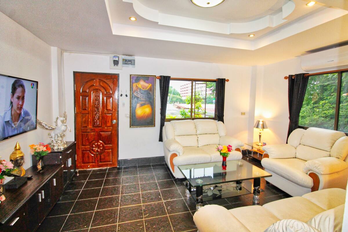 A living room with a sofa, a glass coffee table, and a TV. There is a wooden door and a window with curtains.