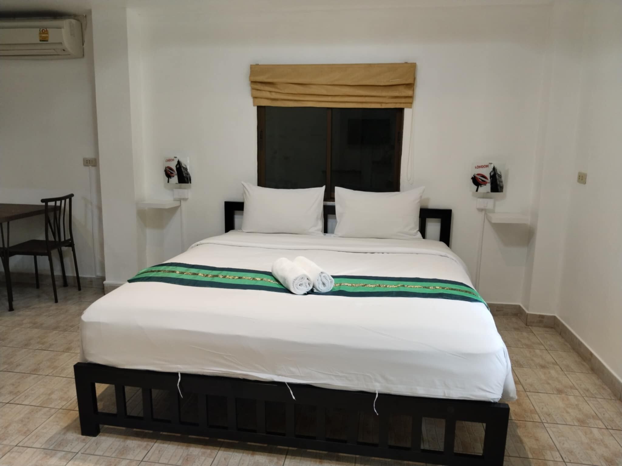 Un lit bien fait, avec une literie blanche et une couverture rayée verte et dorée, une fenêtre agrémentée d’un lambrequin beige, et une petite table avec une chaise placée à côté du lit.